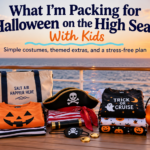 Halloween cruise packing display with orange and black clothing, pirate accessories, pumpkin bucket and travel items arranged on a ship deck at sunset.